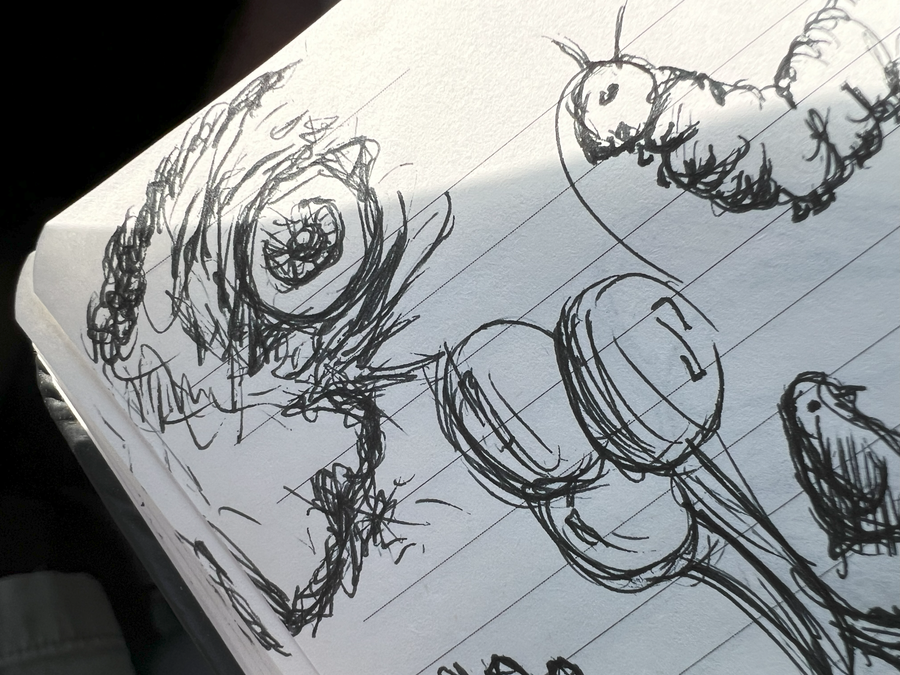 A close-up photograph of a lined sketchbook page, with an exaggerated wide eye drawn in dark, overlapping strokes among loose cartoon shapes and a caterpillar-like doodle.